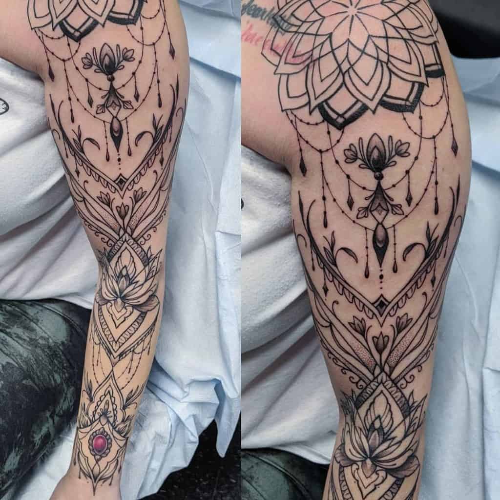 Intricate black ink tattoo featuring mandala patterns, ornamental filigree, lotus flowers, chandelier-style beadwork, and a red gemstone accent