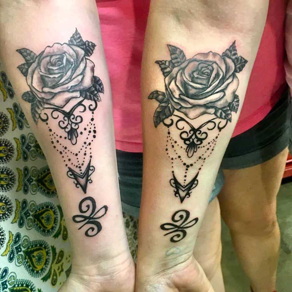 Matching black and grey forearm tattoos featuring realistic roses, ornamental filigree, chandelier-style beadwork, and decorative flourishes