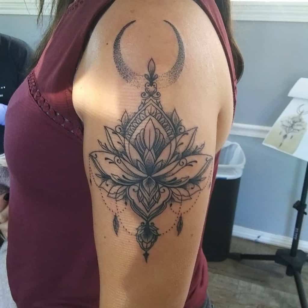 Black and grey tattoo featuring a detailed lotus flower with intricate mandala patterns, a crescent moon above, and chandelier-style teardrop accents