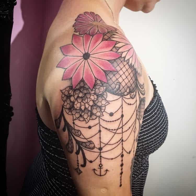 Intricate upper arm tattoo featuring pink flowers, black lace details, cascading chandelier-style beadwork, and a small anchor charm at the bottom