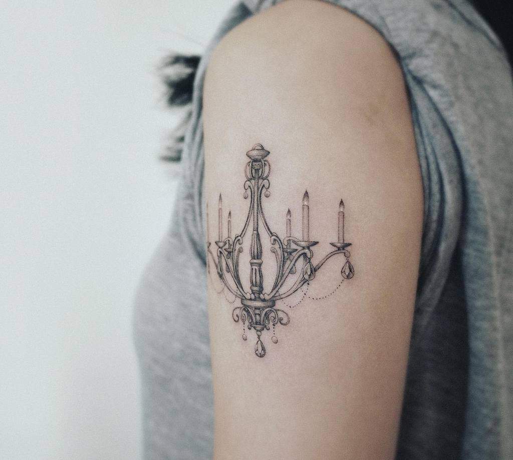 Delicate black and grey upper arm tattoo of a vintage chandelier with six candle lights, intricate metal details, and subtle beaded chains
