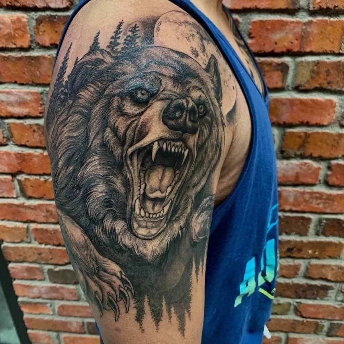 Bear tattoo on upper arm against a moonlit forest backdrop in front of a rustic brick wall