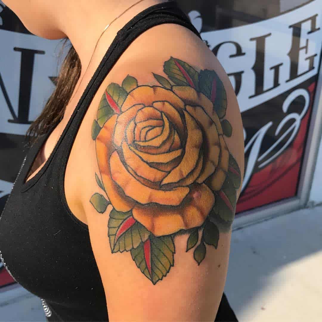 A close-up of a shoulder with a large yellow rose tattoo with green leaves