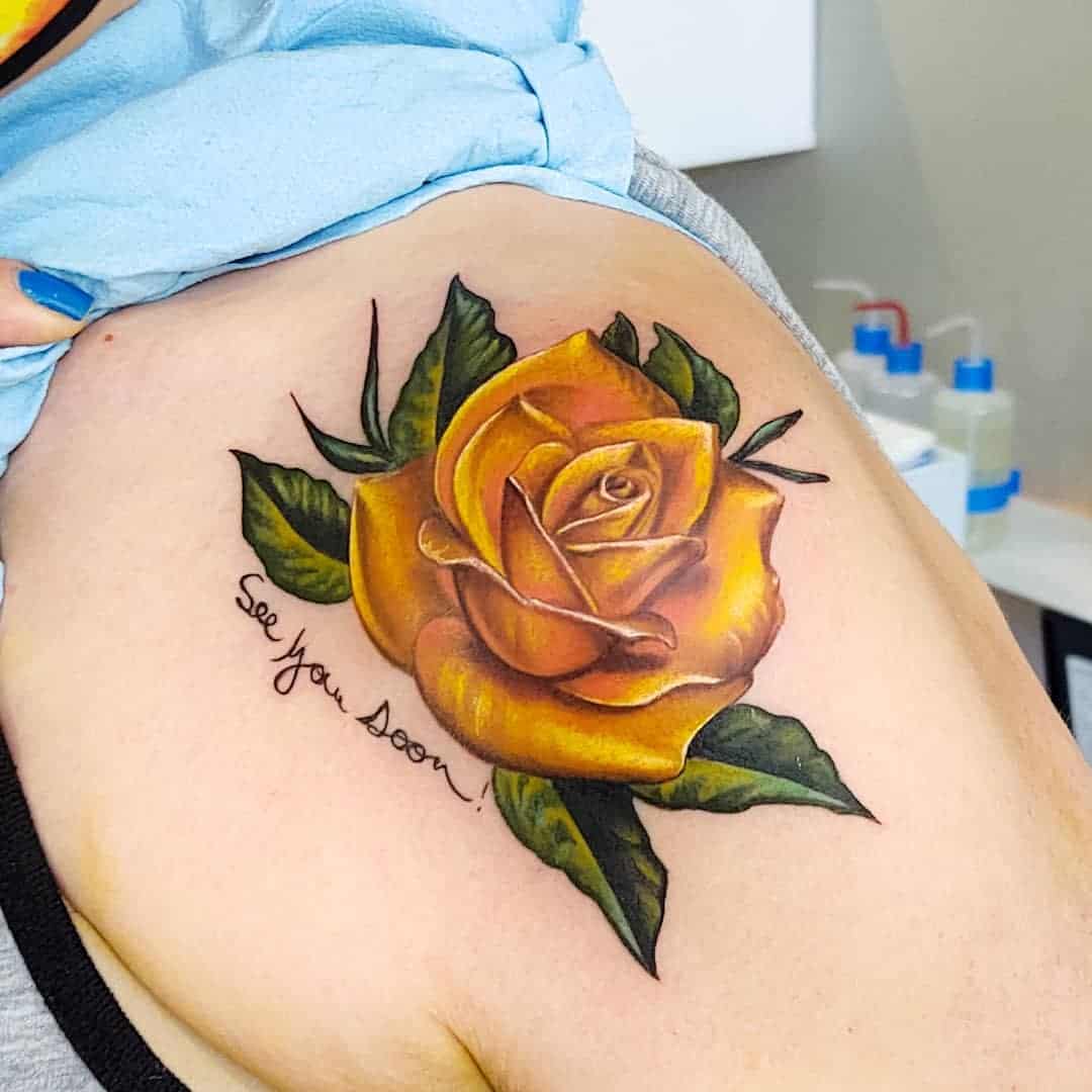 Tattoo of a realistic yellow rose with green leaves and the text "See you Soon!" on an upper arm