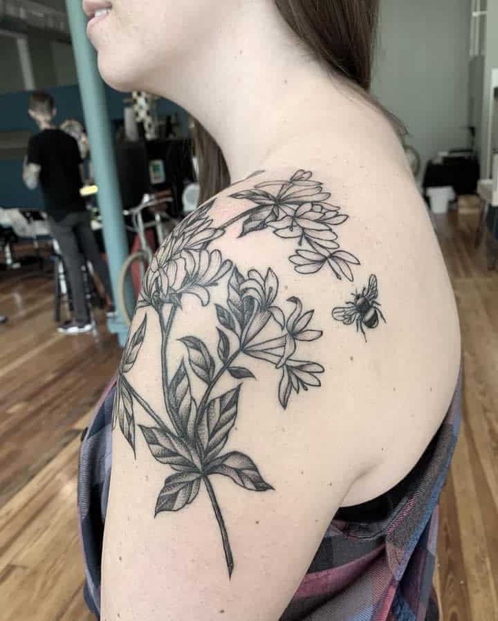 Black ink shoulder tattoo featuring honeysuckle flowers with detailed shading and a hovering bee