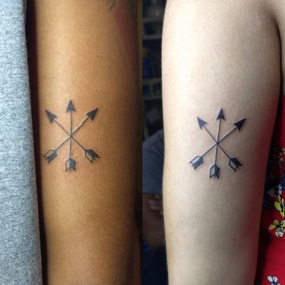Two arms with matching tattoos of crossed arrows pointing outward in a circular pattern