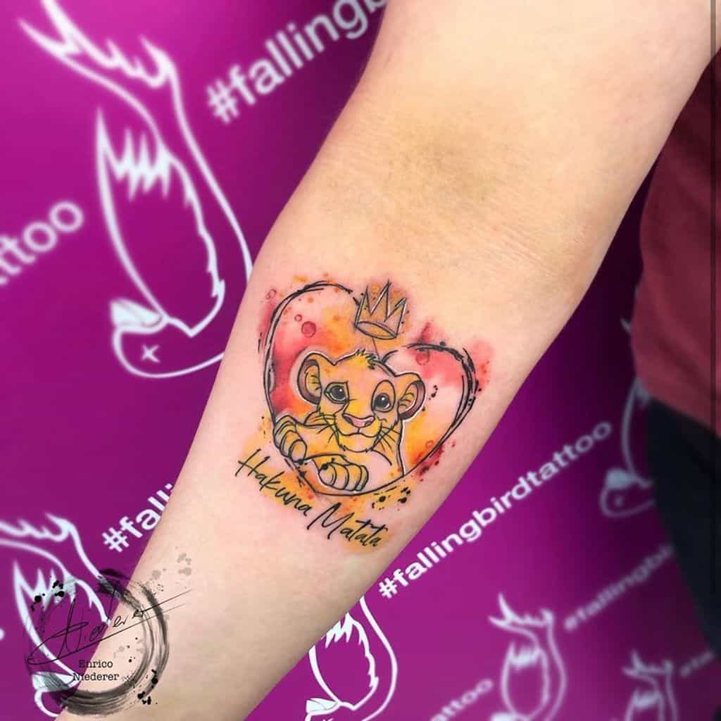 Vibrant tattoo of Simba with a crown inside a heart-shaped outline, surrounded by colorful splashes in orange and red, with the phrase 'Hakuna Matata' below