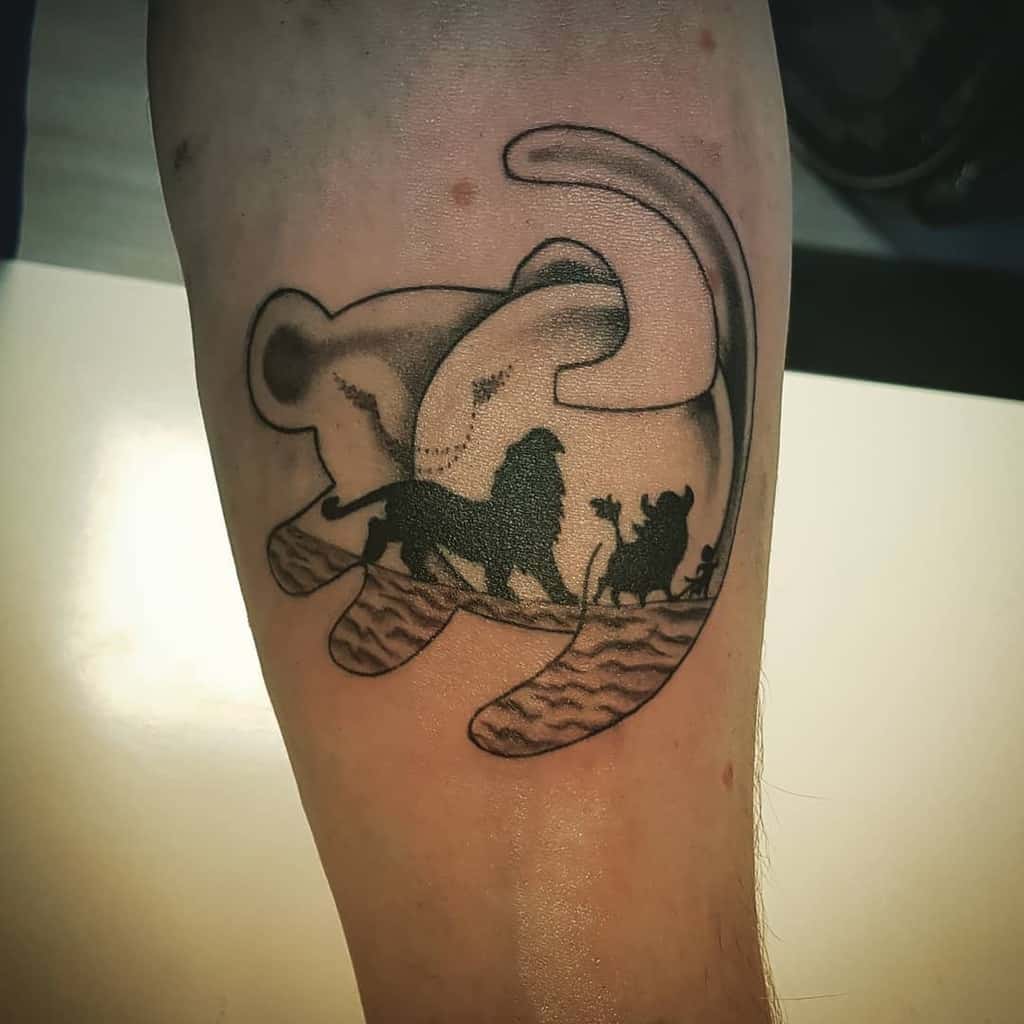 Intricate tattoo featuring a silhouette of Simba, Timon, and Pumbaa walking across the savannah with moon, framed by a Simba's outline in black ink