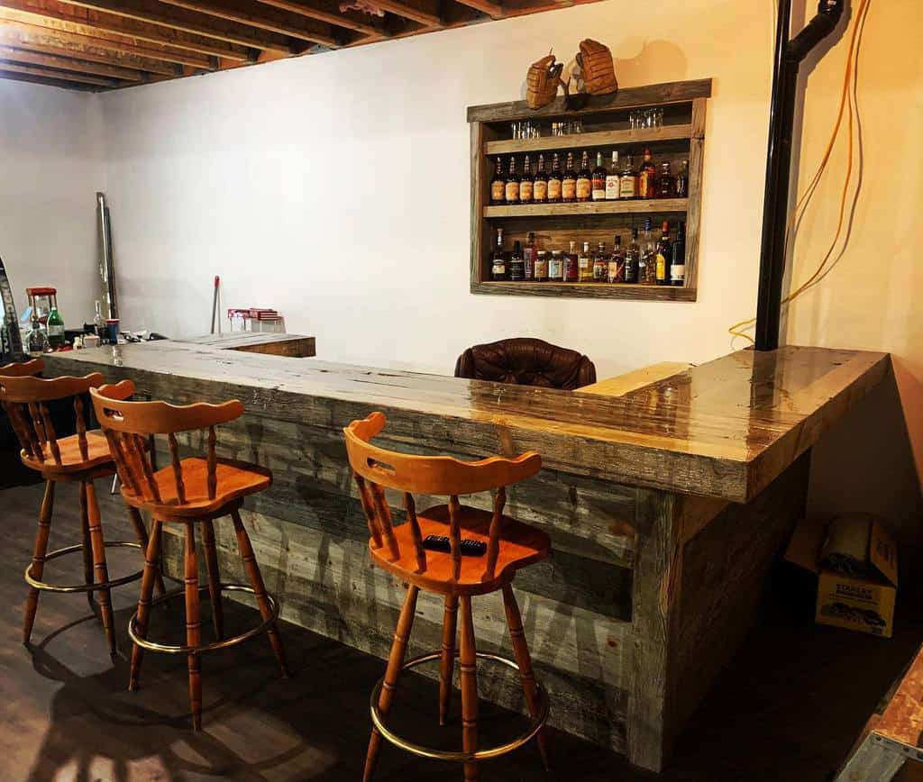Rustic wood basement bar with wooden bar stools and wall-mounted liquor shelf.