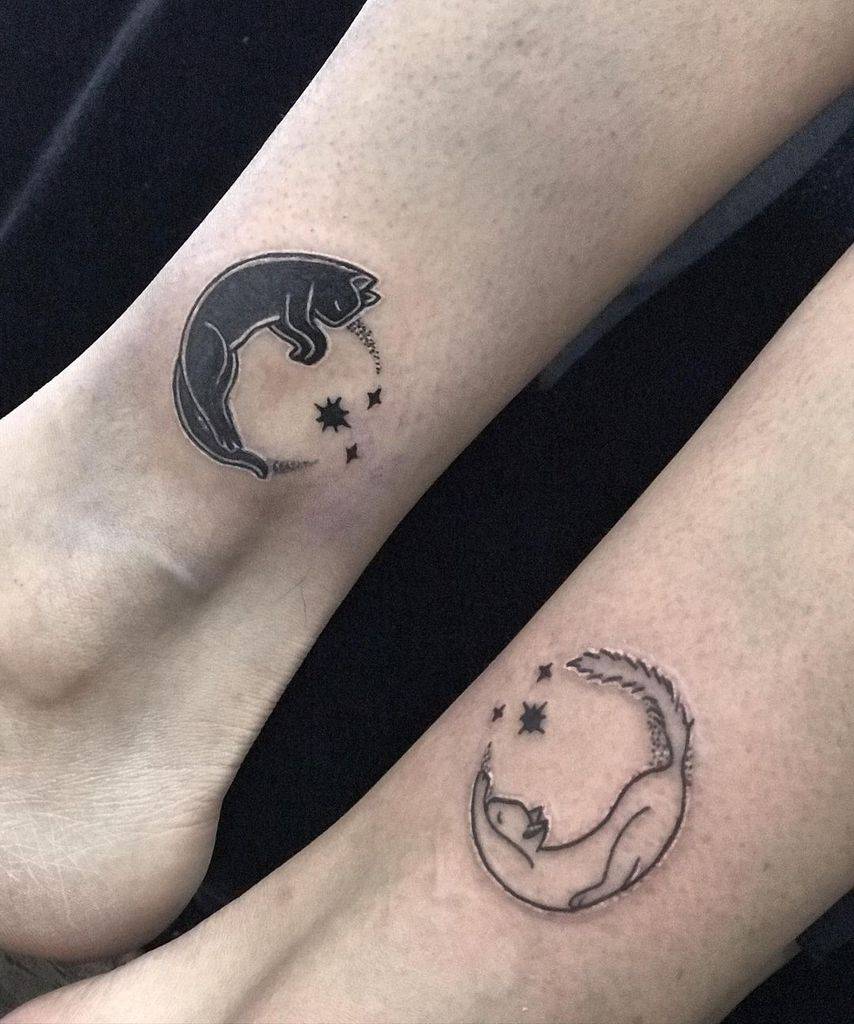 Matching ankle tattoos of cats curled in crescent moon shapes with stars, one in solid black and the other in line art