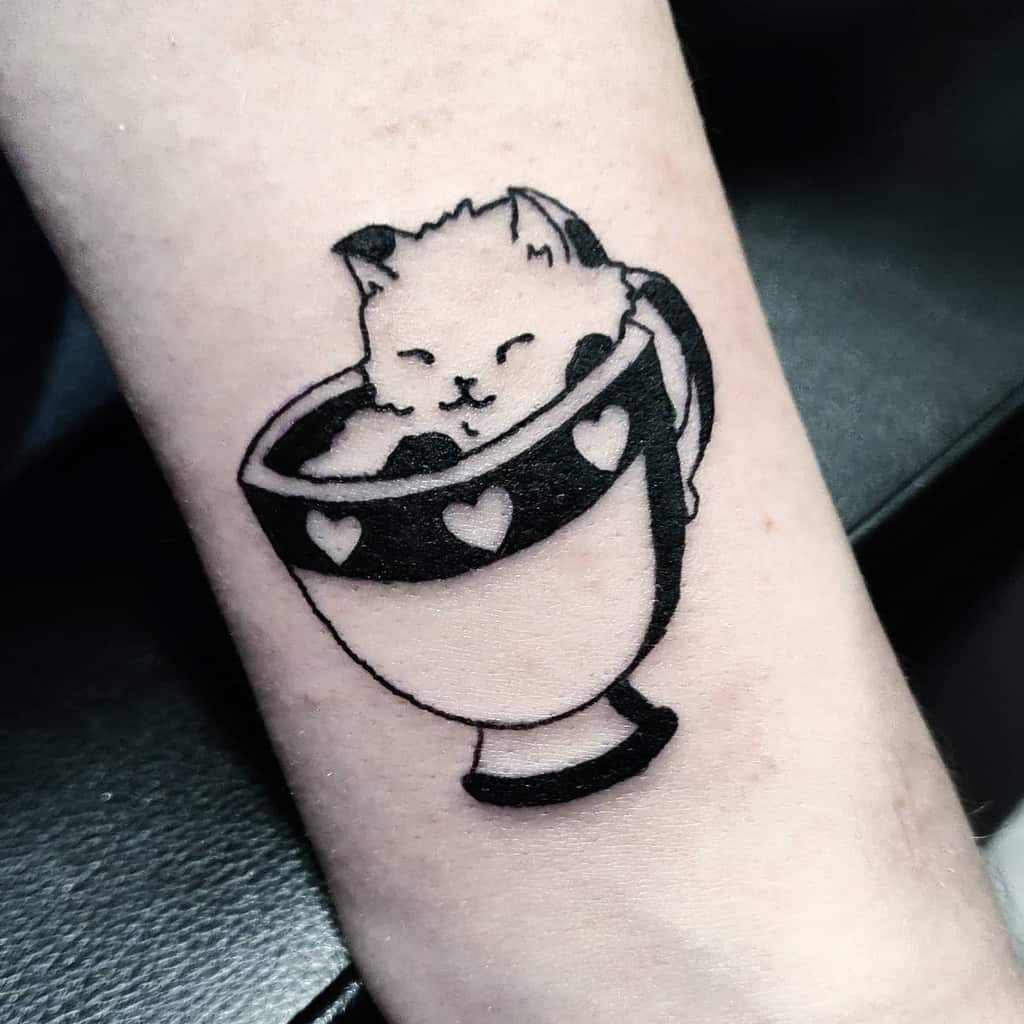 Tattoo of a cat inside a heart-decorated cup on an arm