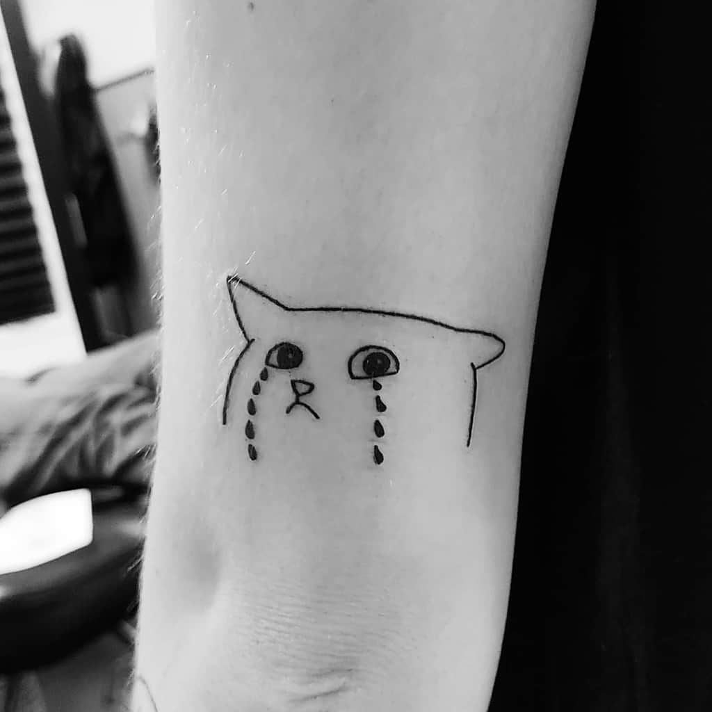 A simple black line tattoo of a sad cat with tears on an arm