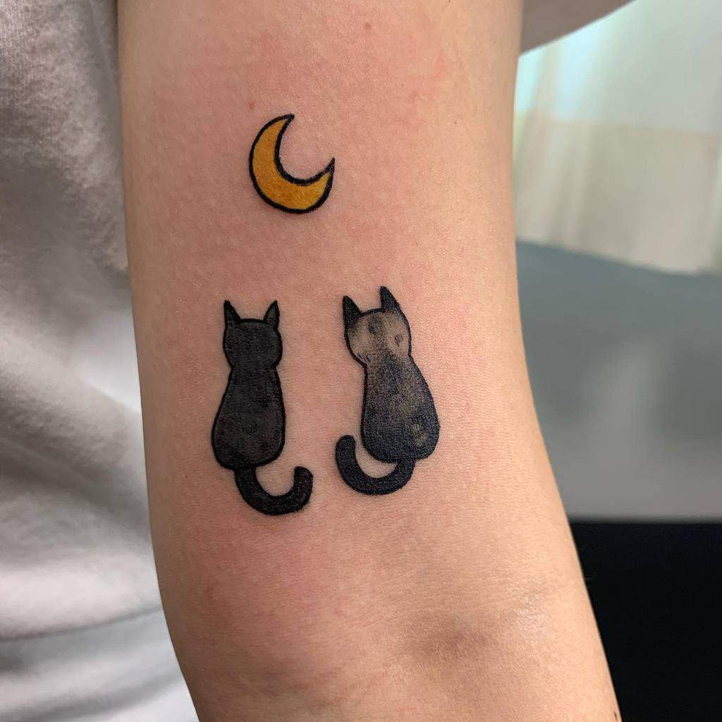 Tattoo of two black cats sitting under a crescent moon on an arm