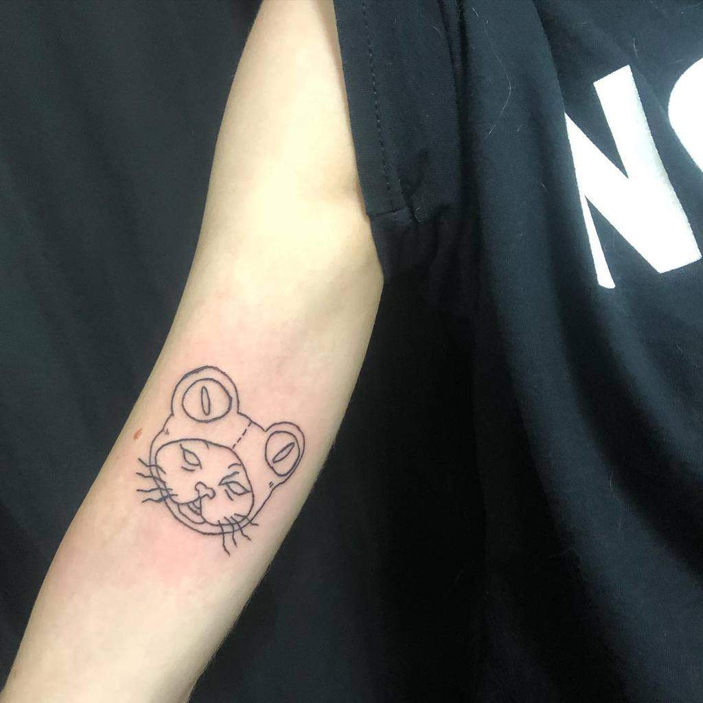 Minimalist tattoo of a cat's face wearing a frog hat, outlined in simple black line art on the upper arm