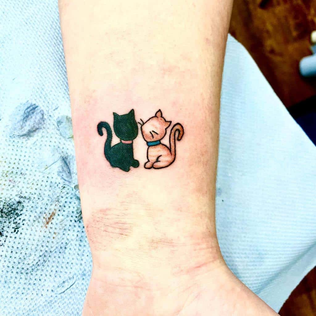 Tattoo of two cats, one black and one orange, sitting next to each other on an arm