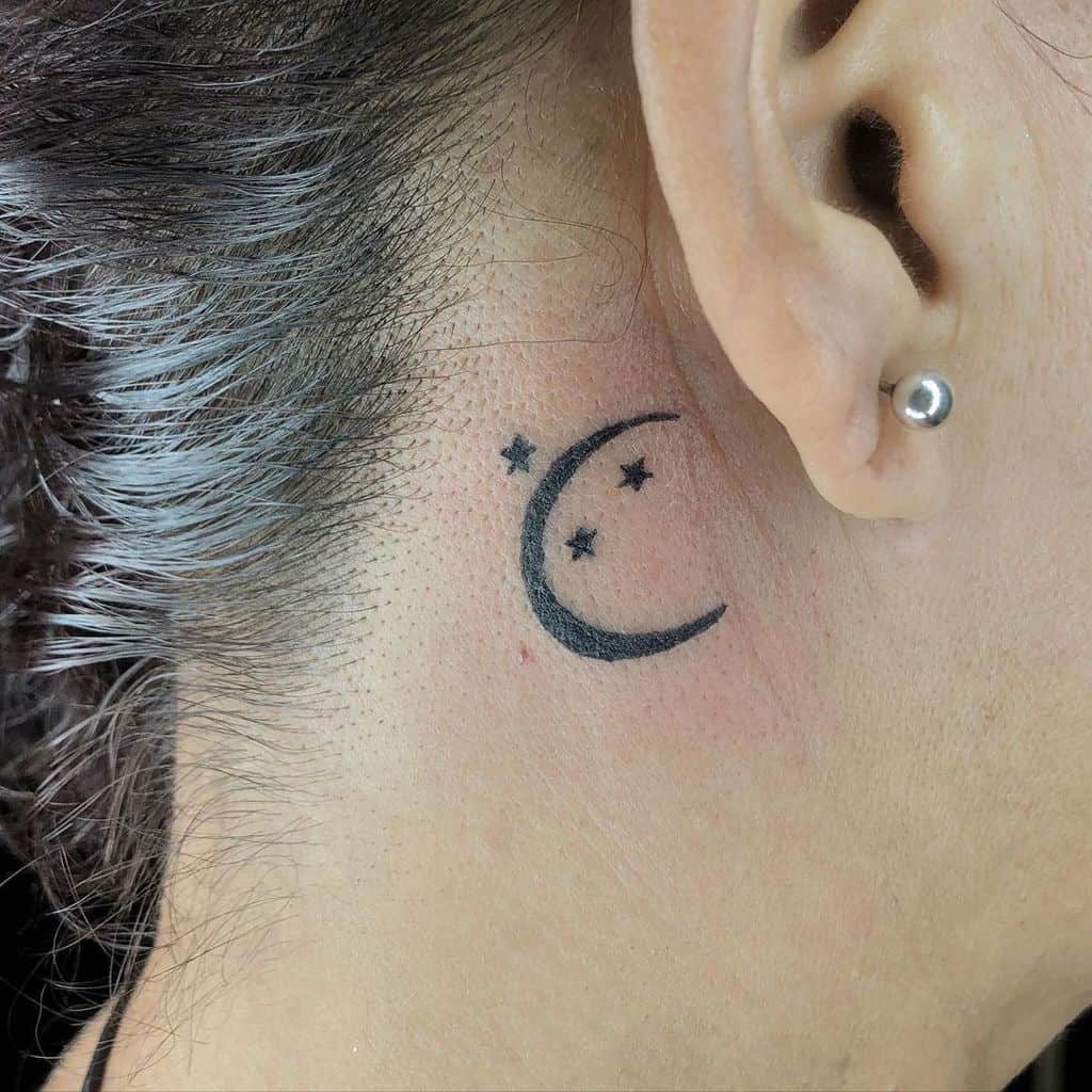 A small crescent moon and stars tattoo behind someone's ear with a delicate ear stud