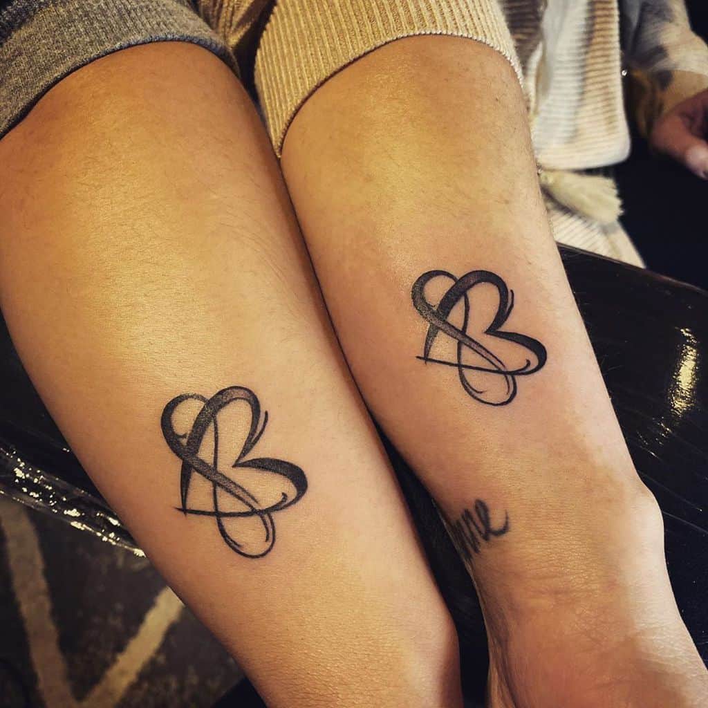 Matching black tattoos on two forearms featuring elegant, cursive infinity symbols intertwined with hearts, symbolizing eternal love