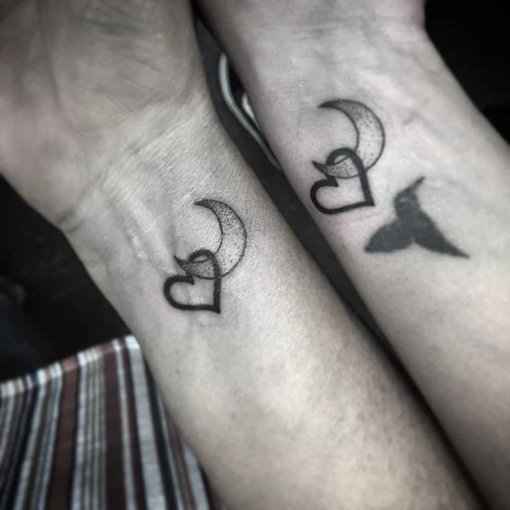 Matching tattoos on two wrists, each featuring a heart combined with a crescent moon and a bird silhouette, symbolizing love and freedom