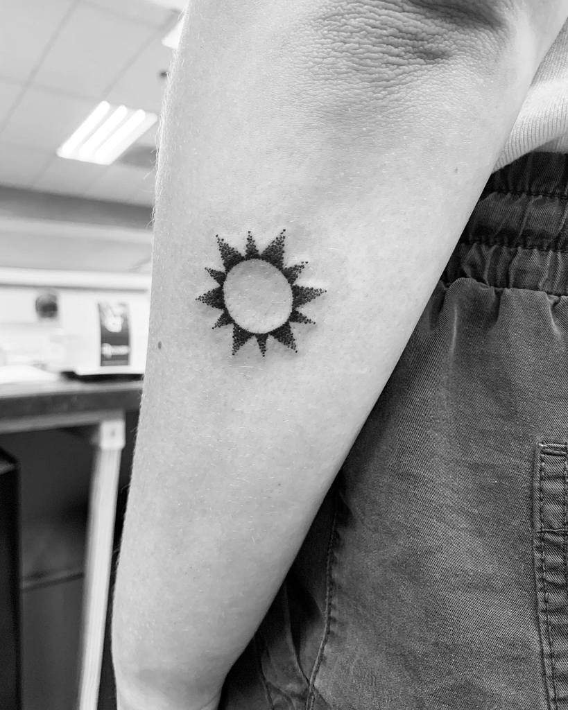 Minimalist black ink sun tattoo with dotwork design, featuring a solid center and dotted rays, placed on the forearm