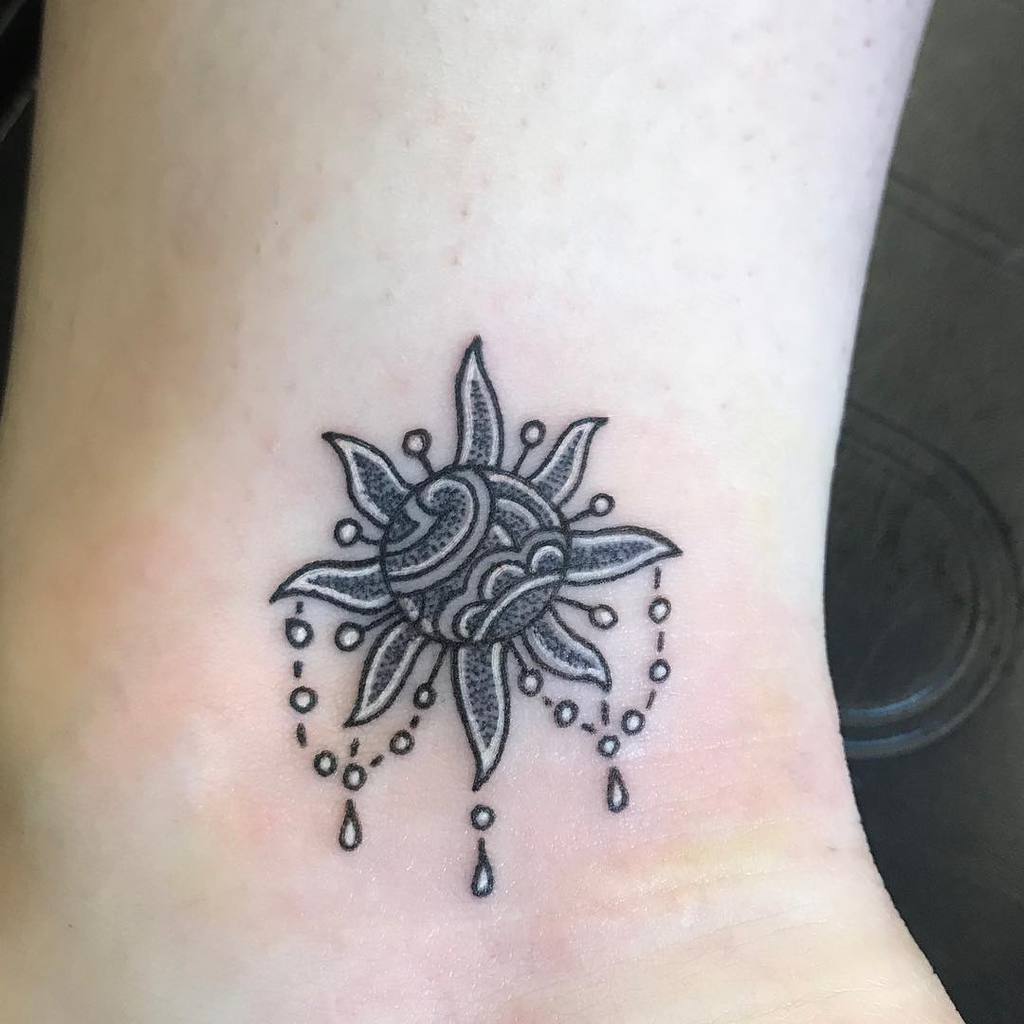 Small ornamental sun and mandala tattoo with detailed patterns, beaded elements, and dripping water-like designs, placed on the ankle