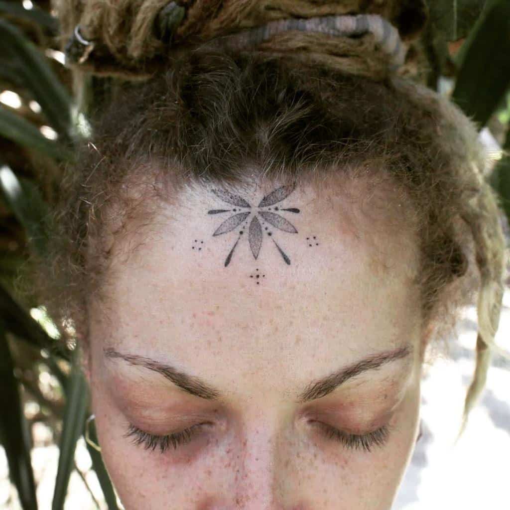 Woman's forehead tattoo featuring a detailed flower-like geometric design with small dotted accents around it