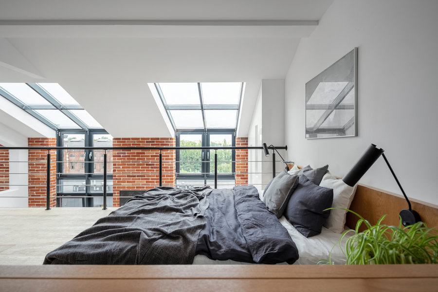 A modern loft bedroom with skylights, a neatly made bed with gray and white linens, brick wall accents, and a railing overlooking a large window