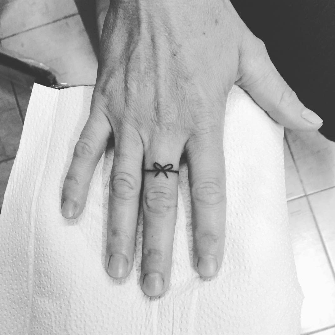 A simple and elegant ring tattoo of a bow, wrapped around the finger, resembling a minimalist ribbon design