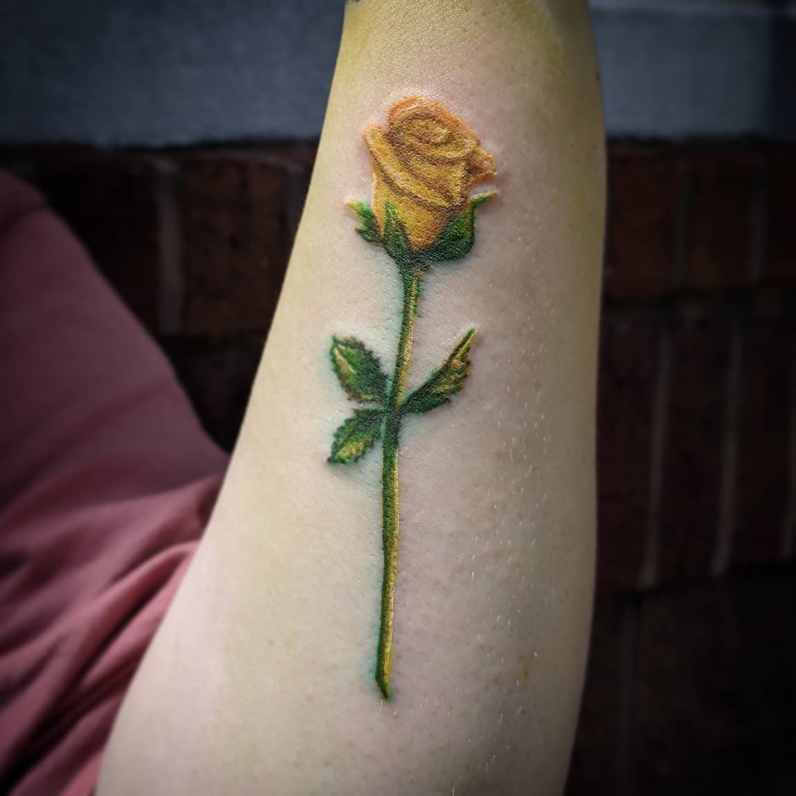 Tattoo of a yellow rose with green stem and leaves on an inner arm