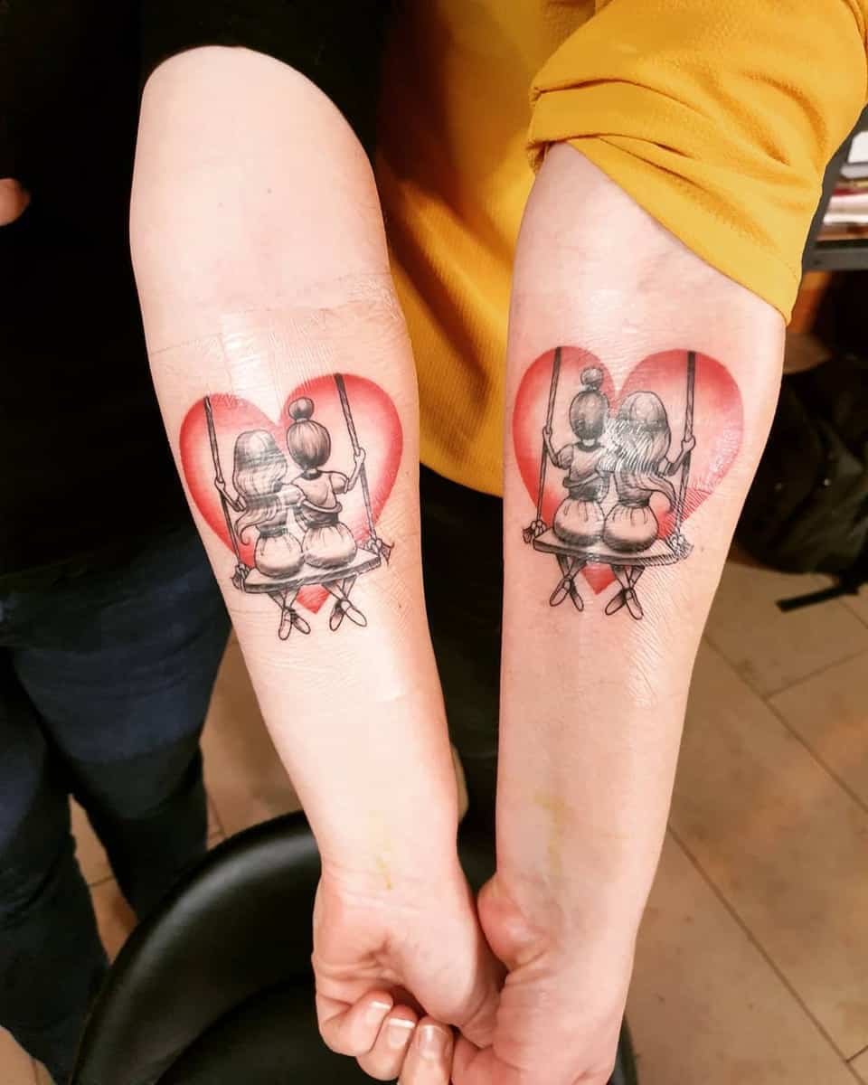 Matching tattoos of two girls on a swing inside a heart on two arms held side by side