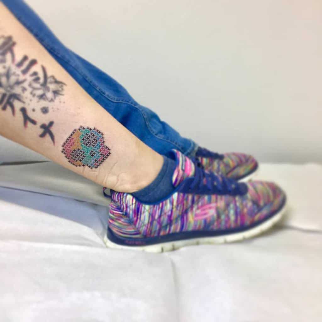 Cross-stitch tattoo of a multicolored skull, designed to mimic embroidered stitches with a pixelated effect for a unique look