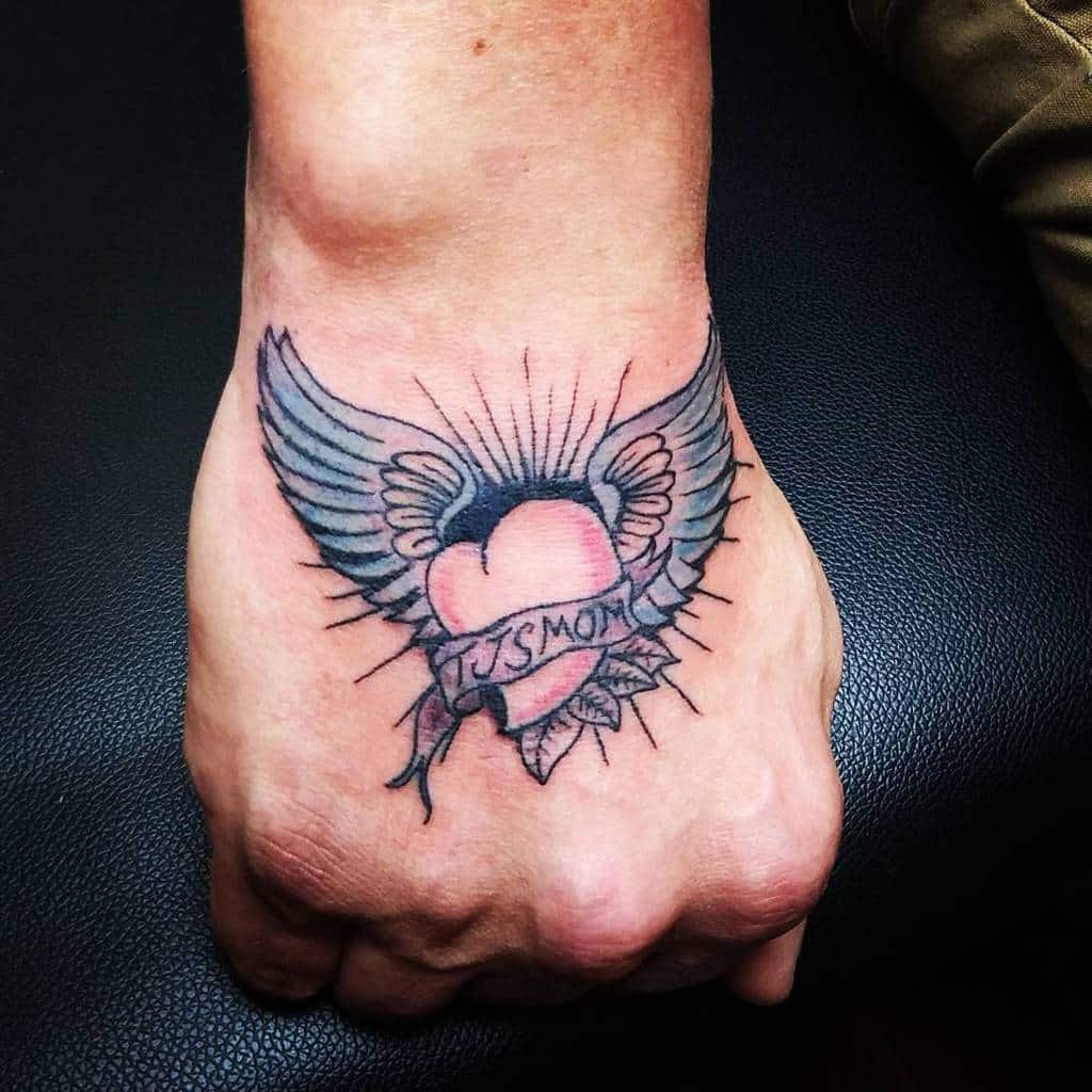 Tattoo of a heart with wings and a banner reading "TJSMOM" on a person's hand