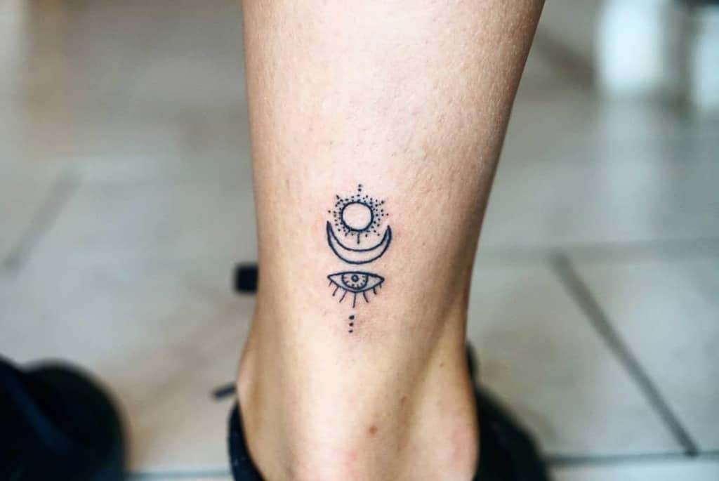 Minimalist tattoo on an ankle, featuring a crescent moon, a dot, and an open eye with lashes