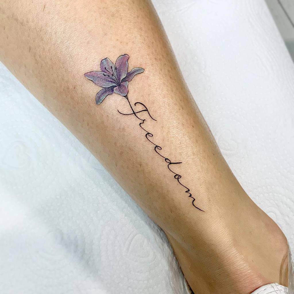 Tattoo of a purple flower with the word "Freedom" written along the stem on a woman's leg