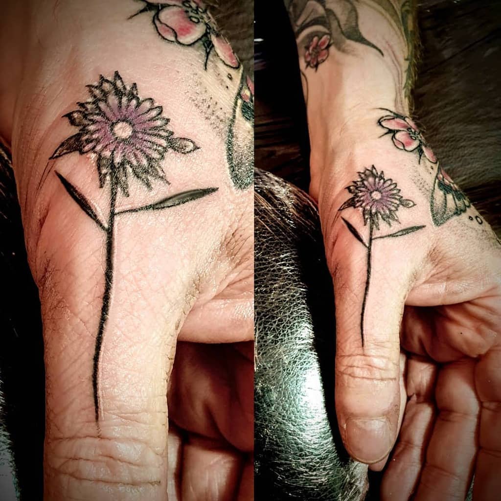 Tattoo of a delicate purple aster flower with a green stem on the side of a person's hand