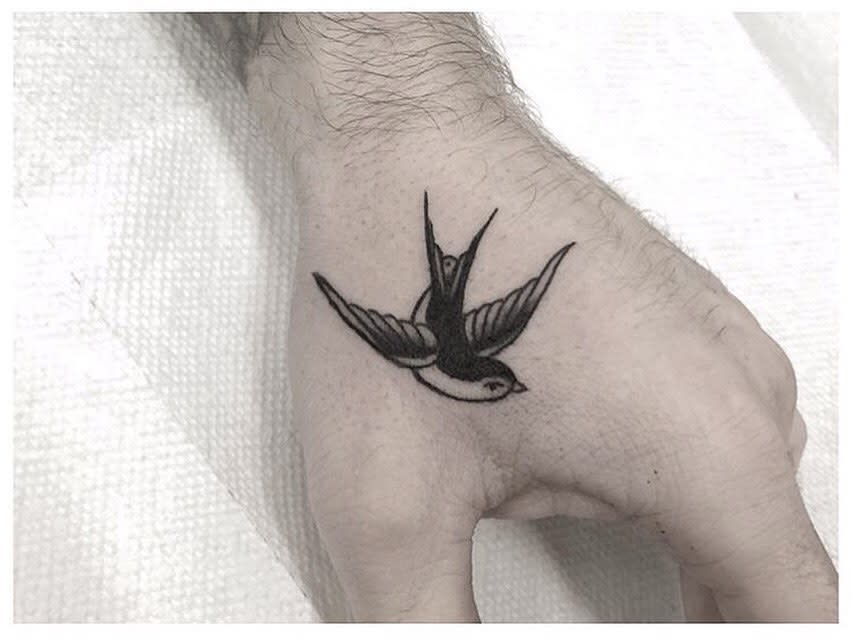Black ink tattoo of a swallow in flight on the skin between thumb and index finger