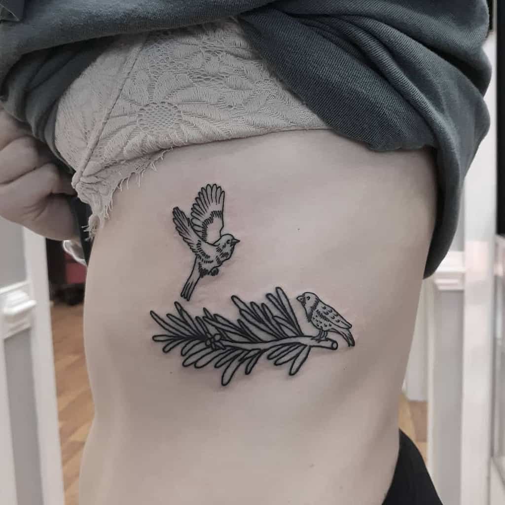 Two birds tattooed on skin, perched on and flying near a pine branch