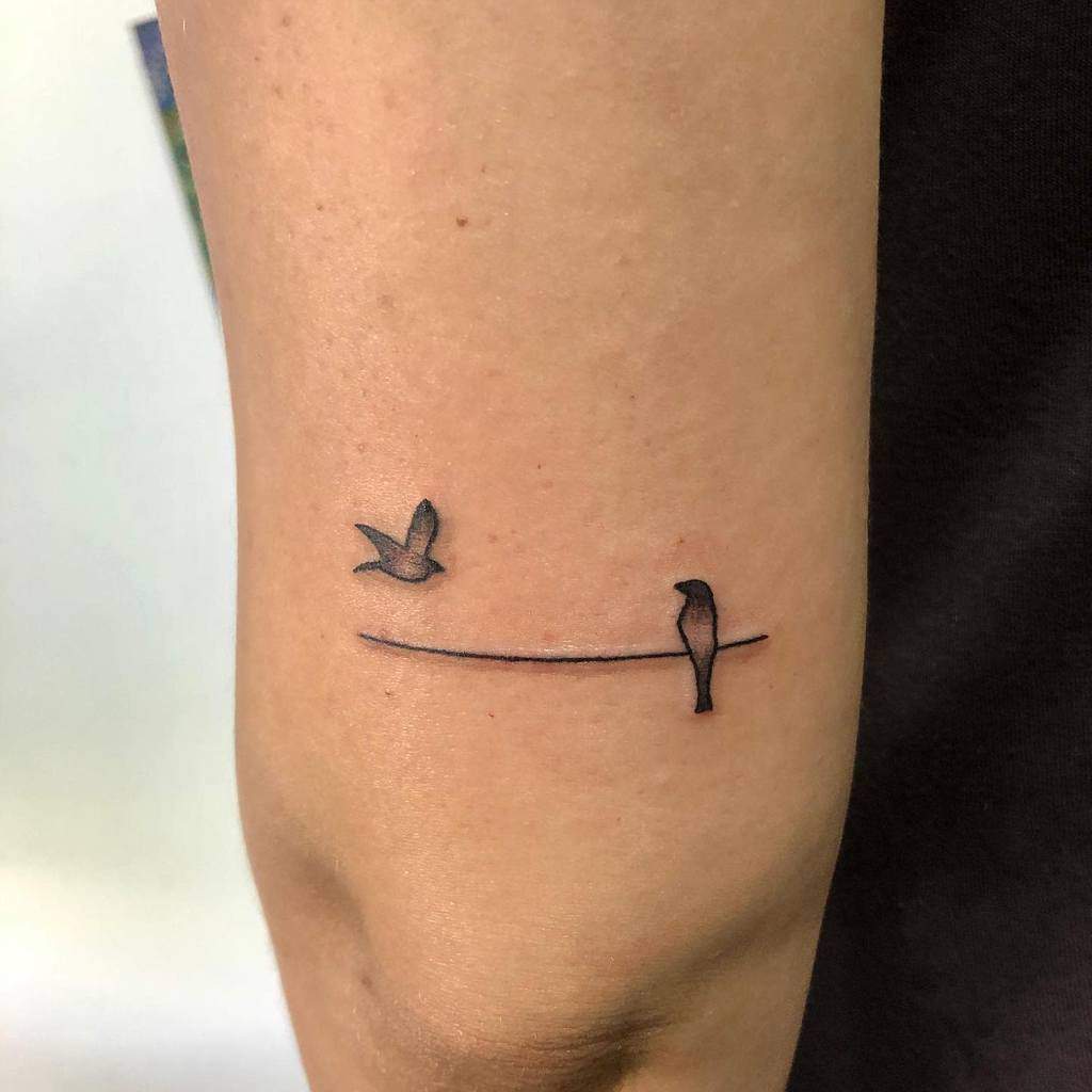 Minimalist tattoo of two birds on an arm, one flying, one perched on a wire