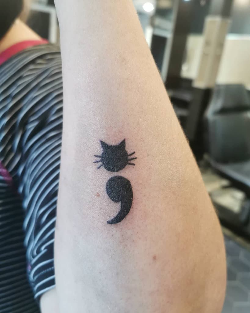 Tattoo of a semicolon with a cat's head on an arm 