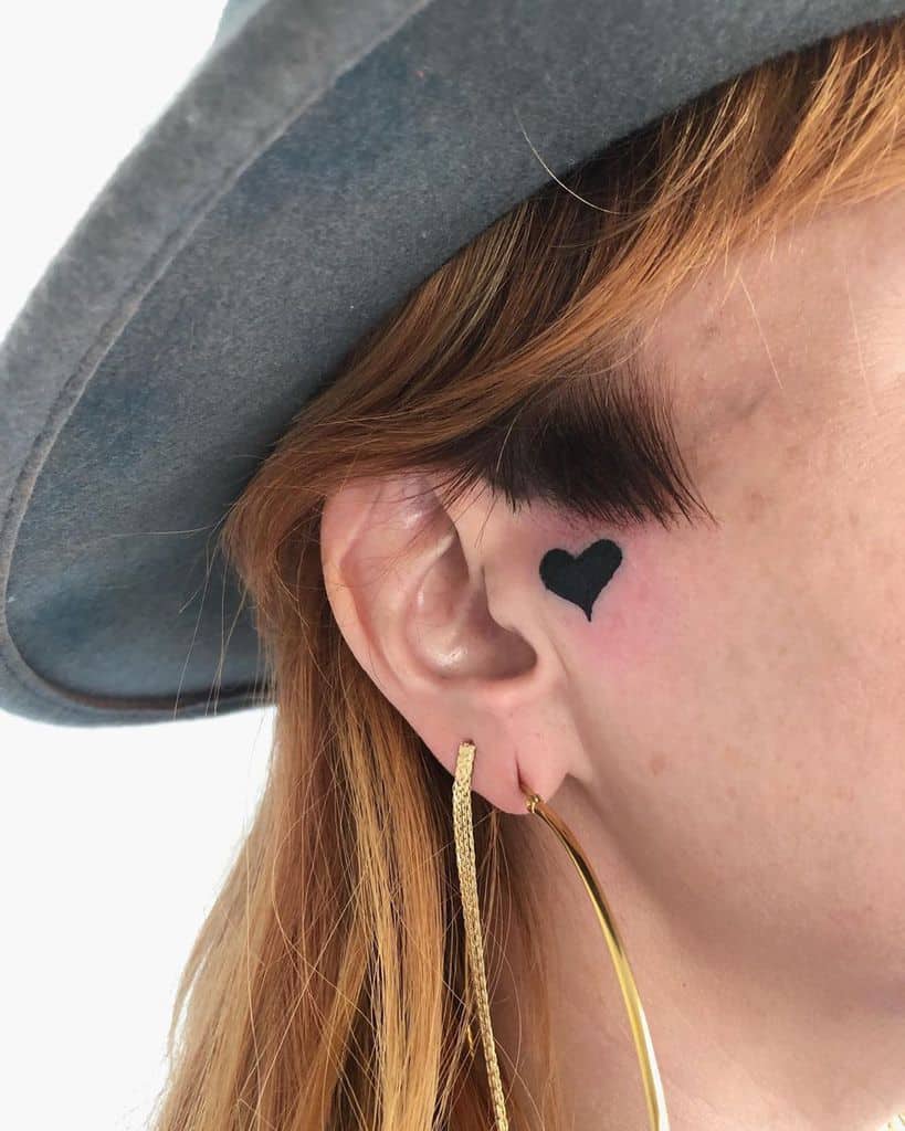 Close-up of a small black heart tattoo just in front of ear