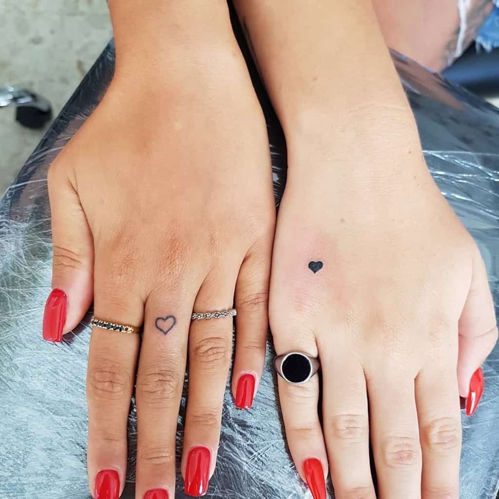Two delicate heart tattoos on hands—one as an outline on a finger and the other as a small solid black heart on the back of the hand