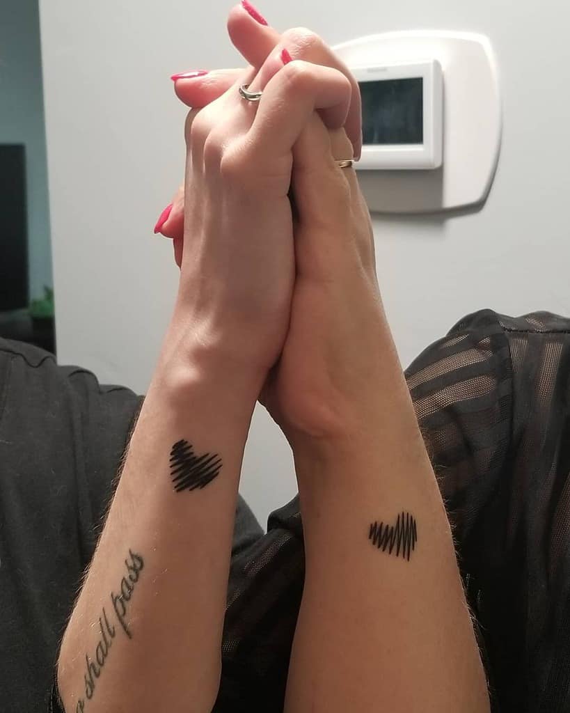 Two forearms with matching black tattoos, each displaying a stylized black heart composed of jagged lines