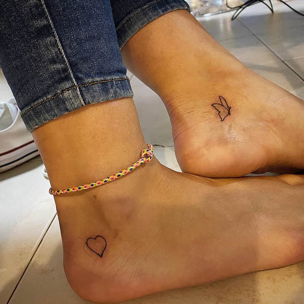 Two ankles close together, each adorned with a small line black tattoo, one of a heart and the other of a butterfly