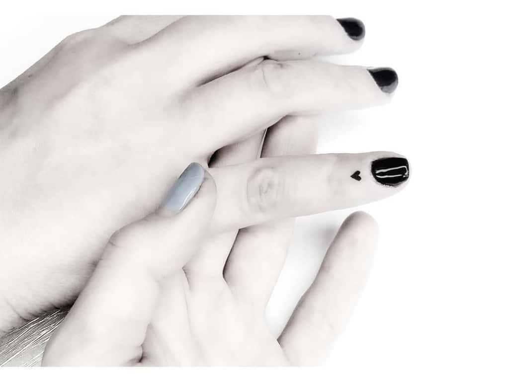 Tiny black heart tattoo on the side of a finger, paired with polished black and pastel nails