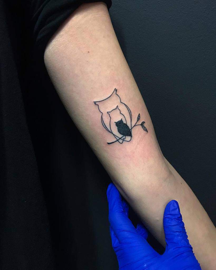 A minimalist black ink tattoo of an owl with a small silhouette of another owl sitting on a branch, placed on the forearm