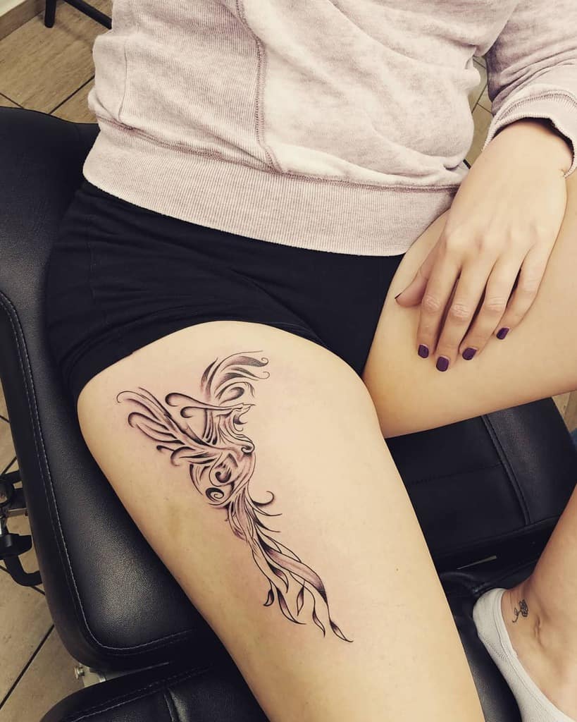 A black and white phoenix tattoo on the thigh, featuring elegant swirls and detailed feathers with intricate line work