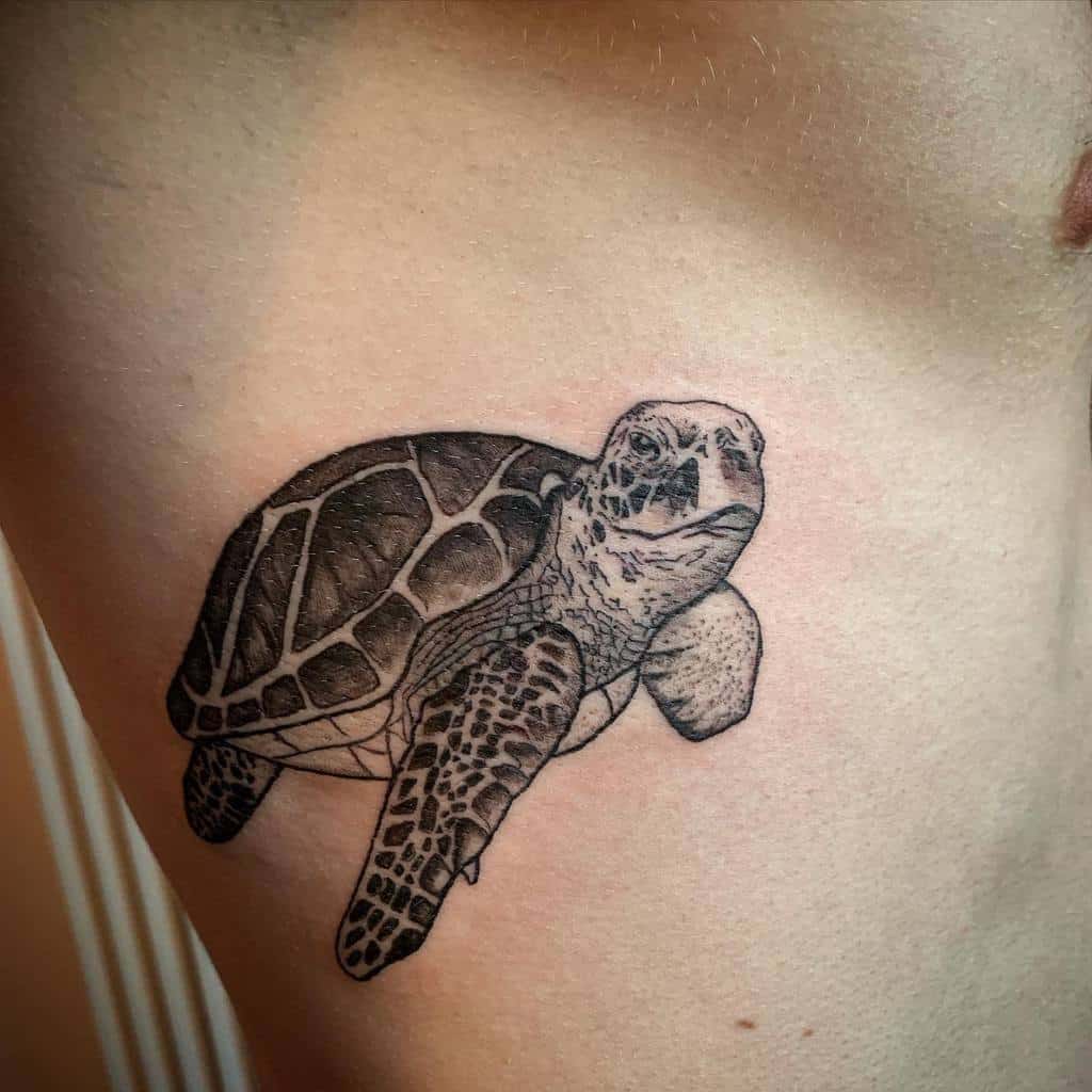 A realistic black and grey tattoo of a turtle, placed on the side of the torso, with intricate detailing on the shell and head