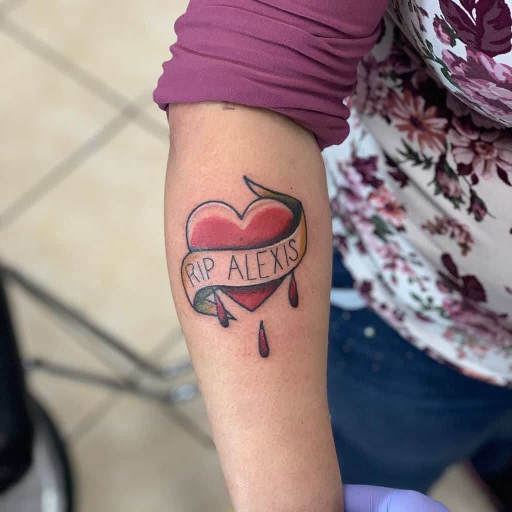 Tattoo of a bleeding heart with "RIP ALEXIS" banner and teardrops on a person's forearm
