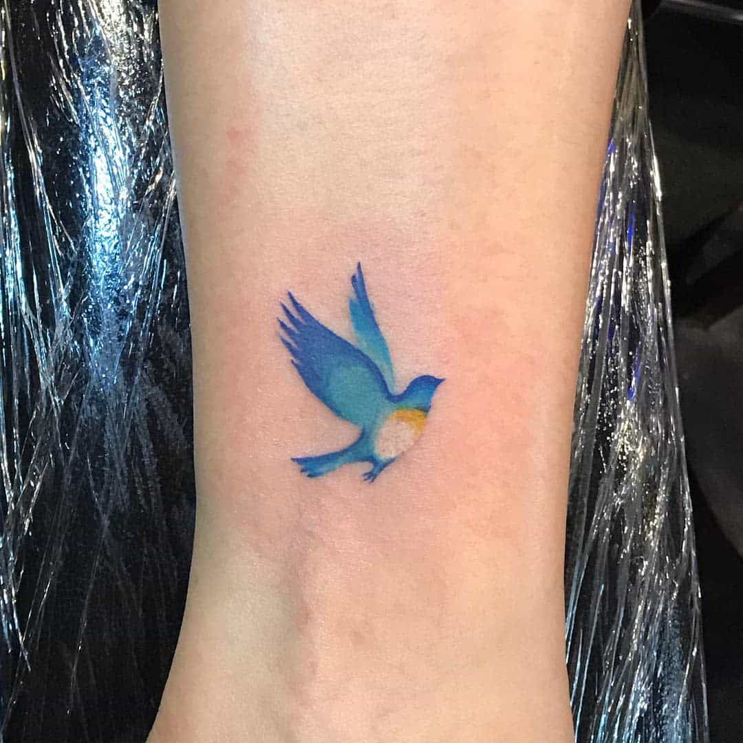 A tattoo of a blue and yellow bird in flight on the inner wrist or lower arm