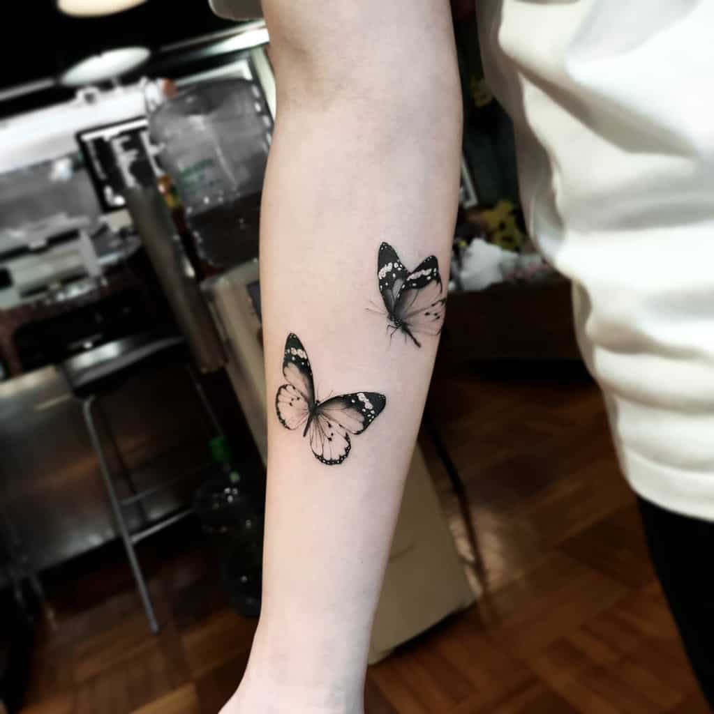 Two black and gray butterflies tattooed on the forearm, featuring fine details and soft shading for a realistic, elegant look.