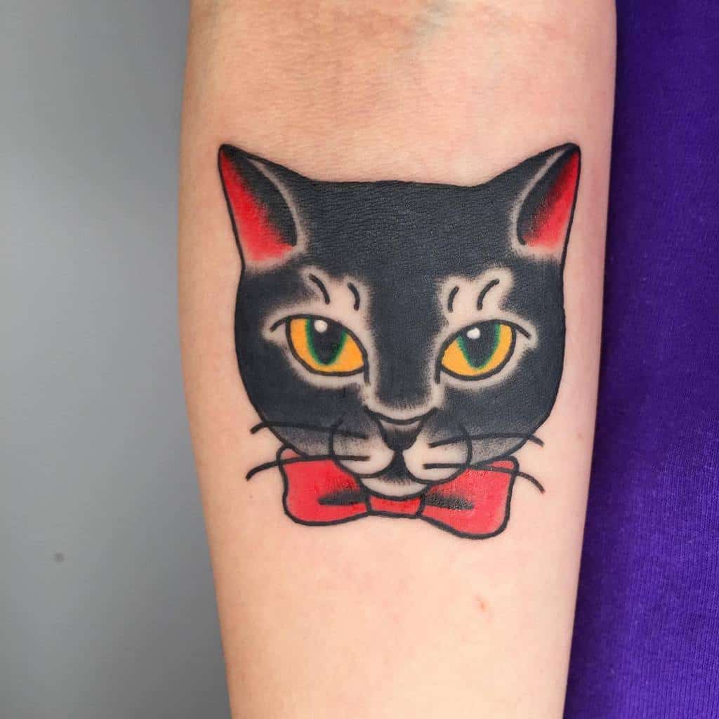 Tattoo of a cat with green eyes and a red bow tie on an arm